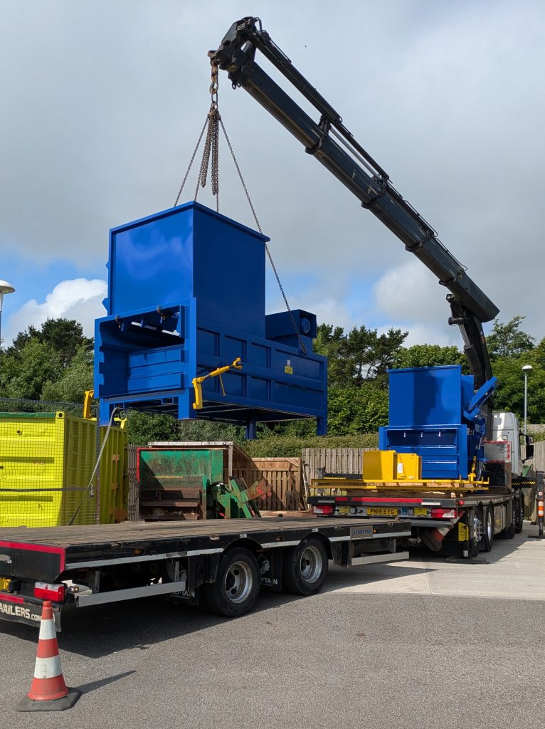 The lorry lifter lifting the large blue waste compactor off of the trailer and into position in the waste compound